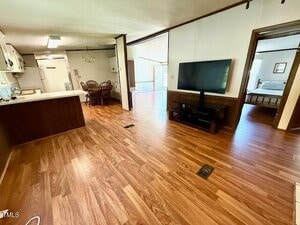 Open living area with wood flooring, featuring a kitchen with white cabinets and appliances, a dining table with chairs, a TV on a stand, and a bedroom visible through an open doorway.