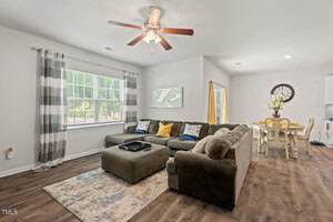 A living room with a sectional sofa, patterned area rug, ceiling fan, and large window with striped curtains. A dining table with chairs and a clock on the wall are visible in an adjacent space.
