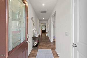 View from an open front door into a well-lit hallway with wood floors, a small table with decor on the left, and rooms visible at the end and side of the hallway. A light-colored rug is placed near the entrance.