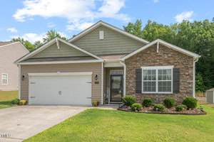Single-story suburban house at 2730 Spring Valley Drive, Creedmoor, featuring a two-car garage, stone and siding exterior, front porch, and landscaped yard with bushes and mulch beds, set against a backdrop of trees and blue sky.