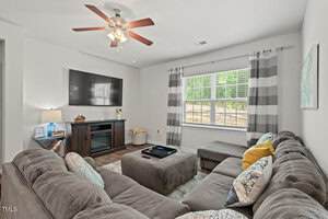 A living room with a large gray sectional sofa, an ottoman, a TV mounted on the wall above a fireplace, a window with striped curtains, a ceiling fan, and decorative pillows on the couch.