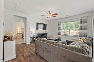 A living room with a gray sectional sofa, a wall-mounted TV above a fireplace, a large window with striped curtains, and wood-style flooring. An open doorway leads to a bedroom with a lit lamp visible.
