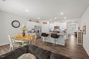 Open concept kitchen and dining area with white cabinets, stainless steel appliances, two barstools at an island, a dining table set for four, wood flooring, and a brown sofa in the foreground.