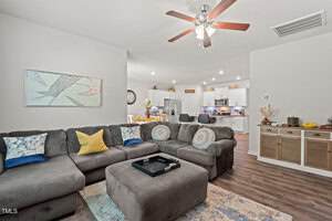 A modern open-concept living room and kitchen with a gray sectional sofa, accent pillows, ottoman, ceiling fan, wall art, and a kitchen area featuring white cabinets and stainless steel appliances.