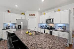 Modern kitchen with white cabinets, stainless steel appliances, granite countertops, and a large island with a sink and seating. Decorative items, small appliances, and utensils are arranged on the counters.