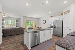 A modern kitchen with white cabinets, stainless steel appliances, and granite countertops, adjacent to a living room with a brown sectional sofa and large windows letting in natural light.