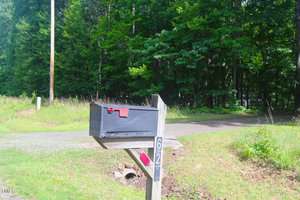 A black mailbox with a red flag is mounted on a wooden post beside a gravel driveway, with the number 627 displayed vertically; green trees and grass are in the background.