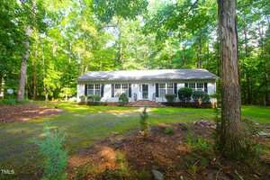 A single-story white house with a gray roof sits in a wooded area at 622 Enon Road, Oxford. The front yard has bushes and a gravel driveway, with tall trees surrounding the house and lawn.