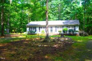 A single-story house with white siding and a gray roof is surrounded by trees. The front yard has a shaded area with sparse grass, rocks, and shrubs near the house. A wooden ramp leads to the front door.