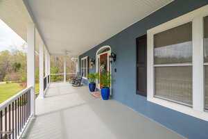 A spacious covered porch with blue walls, a wooden front door, two blue potted plants, black rocker chairs, and a white railing, overlooking a green lawn and trees in the background.