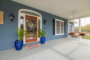 A covered front porch with a wooden door decorated with a wreath, two potted plants in blue planters, and a striped doormat. A porch swing hangs to the right near white railing and a ceiling fan.