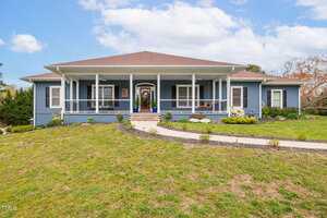 A single-story blue house with a wide front porch and white trim, set on a grassy lawn with landscaped garden beds and a curved concrete walkway leading to the front steps.