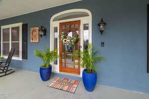 A wooden front door with glass panels features a floral wreath and is flanked by two potted palm plants in blue pots. A striped "Welcome" doormat lies in front, and two black wall lanterns are mounted on either side.