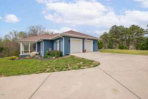 A single-story blue house with a covered porch, attached garage, and light-colored roof sits beside a wide, curved concrete driveway. The lawn is partially green with patches of bare ground and landscaping near the porch.