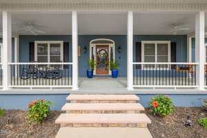 A blue house at 169 Skippers Landing in Manson features a covered front porch with white columns, two ceiling fans, a wooden door with wreath, blue planters, a porch swing on the right, rocking chairs on the left, and steps leading up.
