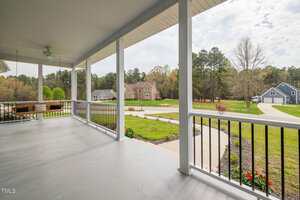 A covered front porch with a ceiling fan, bench swing, and black railing overlooks a landscaped yard, curved driveway, and neighboring houses surrounded by trees.