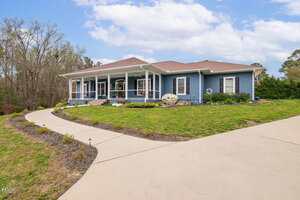 Single-story blue house with white trim, a covered front porch, and a brown roof. The house sits on a grassy lawn with a curved concrete driveway and some landscaping in front. Trees and blue sky are in the background.