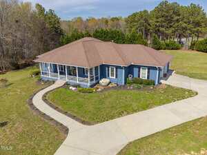 A single-story blue house with a brown roof features a large covered front porch and is surrounded by a curved concrete driveway and landscaped yard. The house is set against a backdrop of trees and grassy areas.
