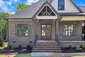 A brick house with a gabled roof has a double wooden front door, steps with metal railings leading to the entrance, two large front windows, and small shrubs along the foundation.