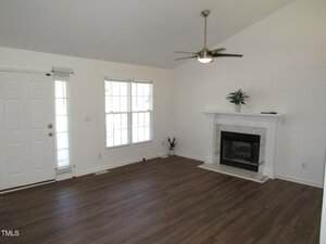 A living room with white walls, wood flooring, a white front door, large windows with blinds, a ceiling fan with light, and a white fireplace with a mantle holding a potted plant.