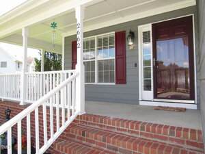 A front porch with brick steps, white railings, gray siding, maroon shutters, a glass storm door, a doormat, a hanging decoration, and the house number 2662 on a column.
