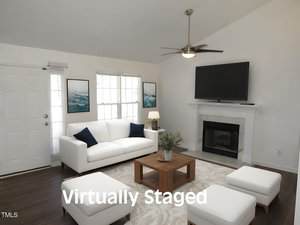 A living room with white walls, a white couch, matching armchair and ottomans, a wooden coffee table, a fireplace, a large TV above the mantel, two windows, and the words "Virtually Staged" overlaid on the image.