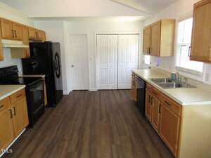 A kitchen with wood cabinets, black appliances, a double sink, white countertops, and wood-patterned flooring. Two white double doors face the kitchen, and a window is above the sink on the right.