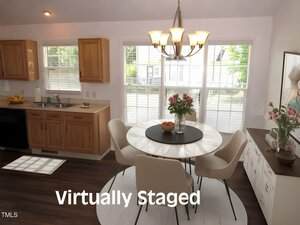 A dining area with a round table and four chairs, a vase of flowers in the center, under a chandelier. Nearby are kitchen cabinets and windows letting in natural light. The text "Virtually Staged" appears at the bottom.