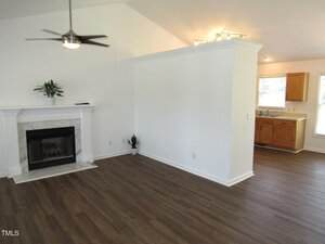 A living area with dark wood flooring, a white fireplace, a ceiling fan, and a plant on the mantel. The space opens to a kitchen with light wood cabinets and white countertops, partially divided by a white partition wall.