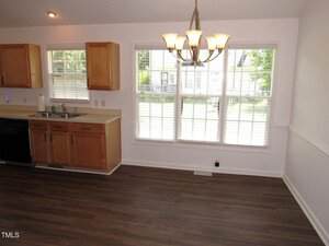 A kitchen and dining area with wood cabinets, a black dishwasher, a sink, large windows with blinds, and a chandelier above a space with wood-look flooring and white walls.