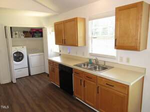 A kitchen with wooden cabinets, a double sink, and a black dishwasher. A laundry area with a washer and dryer is visible in an open closet to the left. The floor is brown wood and there is a window above the sink.