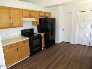 Kitchen with light wood cabinets, black appliances including a stove and refrigerator, white countertops, wood-look flooring, and white doors, one of which appears to be a closet or pantry.