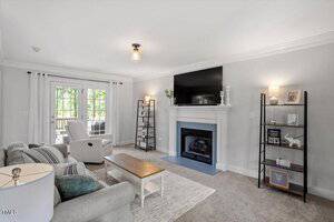 A living room with a gray sofa, white armchair, wooden coffee table, fireplace with a TV above it, two shelving units with decor, and French doors letting in natural light. The walls are light gray and the carpet is beige.