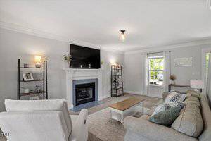 A living room with a gray sofa, white armchair, wooden coffee table, and fireplace with a TV mounted above. Shelving units stand on either side of the fireplace. A window with white curtains lets in natural light.
