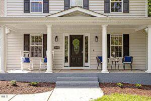 A two-story house with light gray siding and black shutters features a covered front porch with white columns, two rocking chairs, a small table, and navy blue pillows on the chairs. The front door is black with a glass panel.