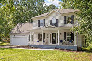 Two-story white house with black shutters, a covered front porch with chairs, and a two-car garage. The yard has grass and trees, and the house is surrounded by greenery under a partly cloudy sky.