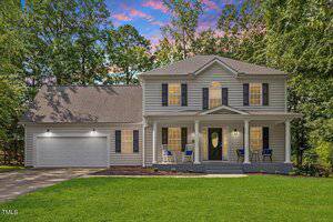 Charming two-story house at 1990 Ferbow Street, Creedmoor, with gray siding and dark shutters, a front porch with two chairs and a table, double garage on the left, well-kept lawn, trees behind the home, and a colorful sunset sky.