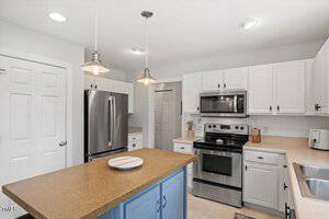 A modern kitchen with white cabinets, stainless steel appliances, a beige countertop, a kitchen island with a brown surface and blue base, and pendant lights hanging above the island.