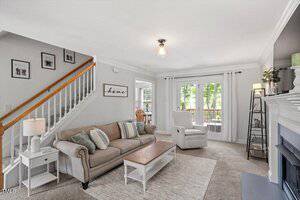 A neutral-toned living room with a beige sofa, striped and solid pillows, a white armchair, a wooden coffee table, and a fireplace. A staircase with wooden handrail and framed art is on the left; French doors open to a deck.