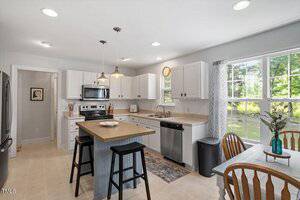 A bright kitchen with white cabinets, stainless steel appliances, a center island with two stools, a dining table with chairs by a window, and light-colored countertops. Sunlight streams in through large windows.
