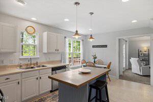A modern kitchen with white cabinets, stainless steel appliances, and an island with barstools. Large windows let in natural light over a dining area with a wooden table and chairs. A living room is visible through an open doorway.