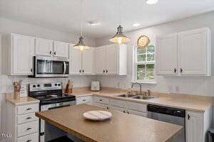 A kitchen with white cabinets, stainless steel appliances, a double sink under a window, beige countertops, two pendant lights, and a round plate on a center island.
