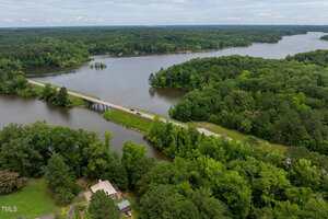 A long, narrow bridge crosses over a river surrounded by dense green forest, with several houses partially visible in the foreground and wooded shoreline extending into the distance.