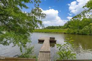 A wooden dock extends over a calm lake surrounded by green trees and shrubs under a partly cloudy sky. The water is still, and the dock has a small platform at the end.