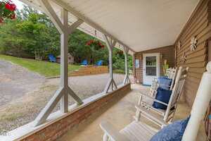A covered porch with white rocking chairs and blue cushions faces a gravel driveway and a grassy area with Adirondack chairs. Hanging flower baskets and outdoor decorations are visible.