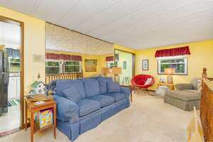 A living room with yellow walls, a blue sofa, an upholstered armchair with a red cushion, a side table with a lamp, and large windows. A kitchen area is visible through an open doorway on the left.