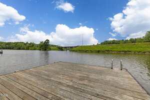 A wooden dock extends over a calm river with green grassy banks; a bridge and utility poles are visible in the background under a blue sky with scattered clouds.
