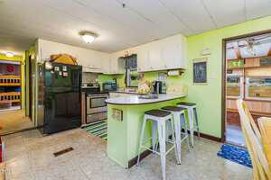 A small kitchen with light green walls, white upper cabinets, a black refrigerator, stainless steel stove, and a breakfast bar with three white stools. A hallway and another room are visible in the background.