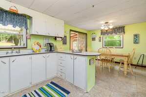 A kitchen with white cabinets, a coffee maker, and a striped rug leads into a dining area with a wooden table and chairs, green walls, and large windows letting in natural light.