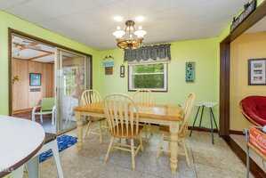 A dining area with a wooden table and four chairs, light green walls, a window with a patterned valance, a ceiling light fixture, and sliding glass doors leading to a sunroom.